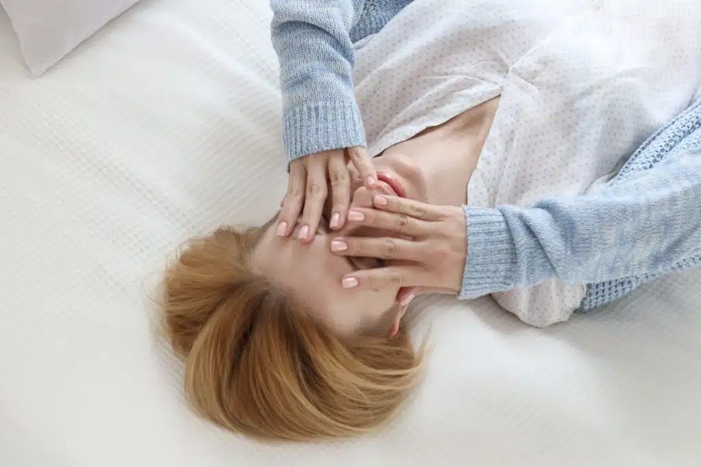 Woman lying on a white bed, covering her face with both hands